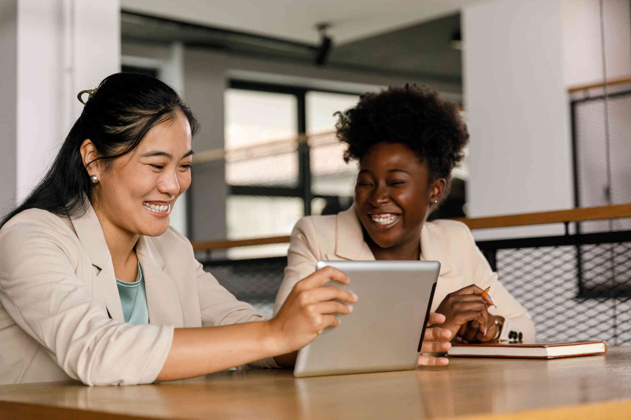 Two women smile widely as they sit at a table in business formal attire and look at a tablet one of them is holding in their hands.